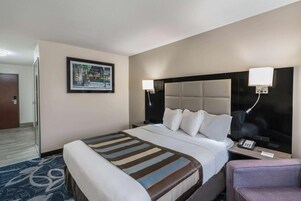 Habitación de hotel con una cama grande, mesitas de noche, un escritorio, una silla, un cuadro en la pared y una puerta.