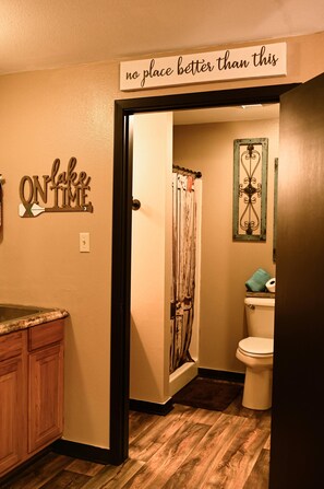 A bathroom with a wooden floor, a toilet, a shower curtain, and a sign that reads 'no place better than this.' - #3 Uncertain Oasis-Uncertain Lodge and Inn (Karnack)