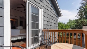 An open sliding door leading to a bedroom with a bed and a ceiling fan. A wooden deck with a table and chairs is visible.