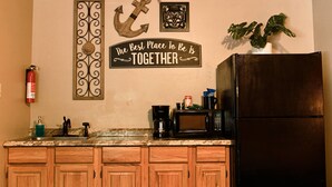 Una cocina con armarios de madera, un refrigerador negro, un microondas y una cafetera en la encimera. Hay un extintor en la pared y un letrero decorativo que dice "The Best Place To Be Together".