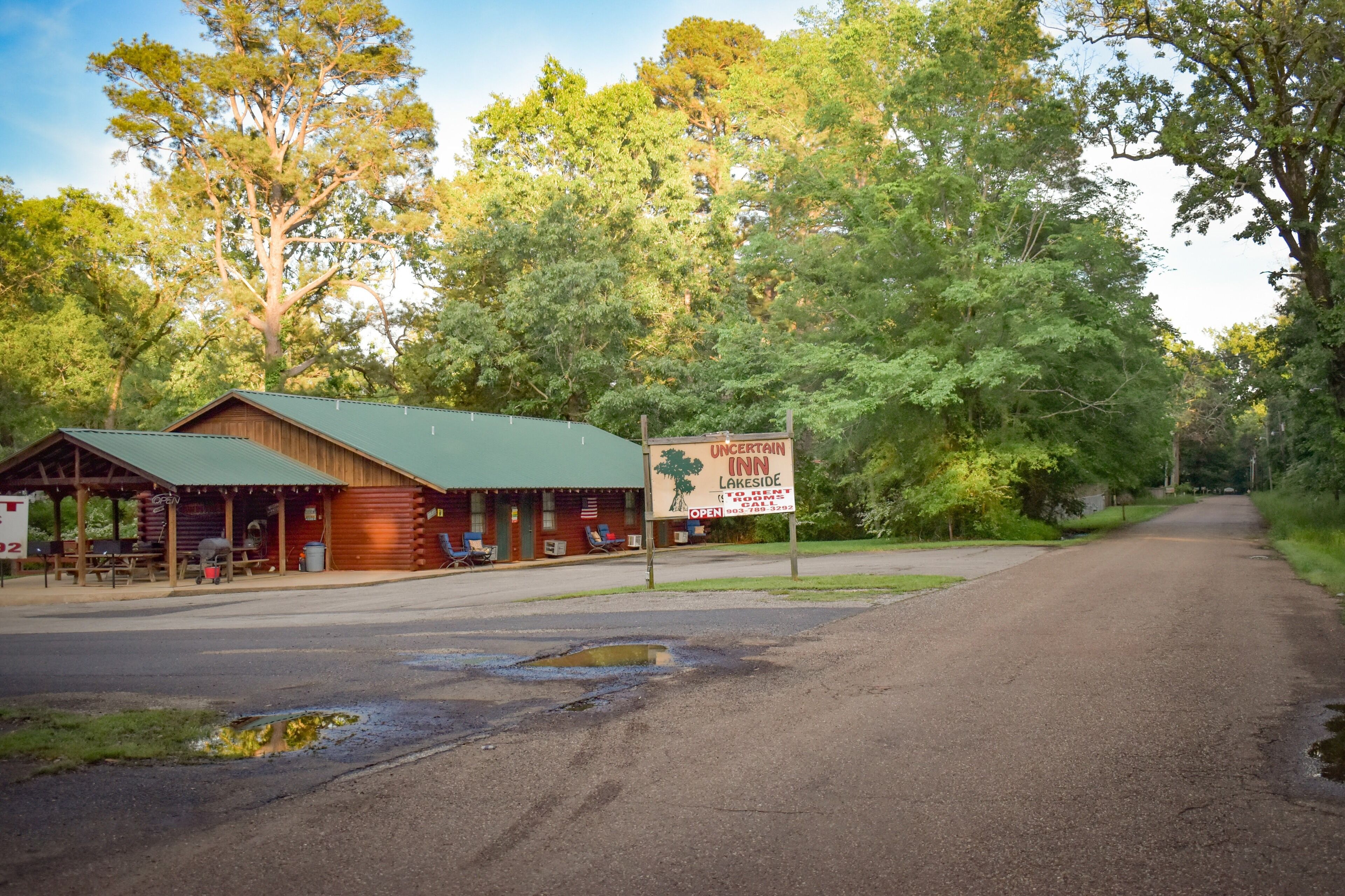 A rustic lodge with a green roof and wooden exterior, a signboard advertising 'On Certain Inn', and a road leading into a wooded area.