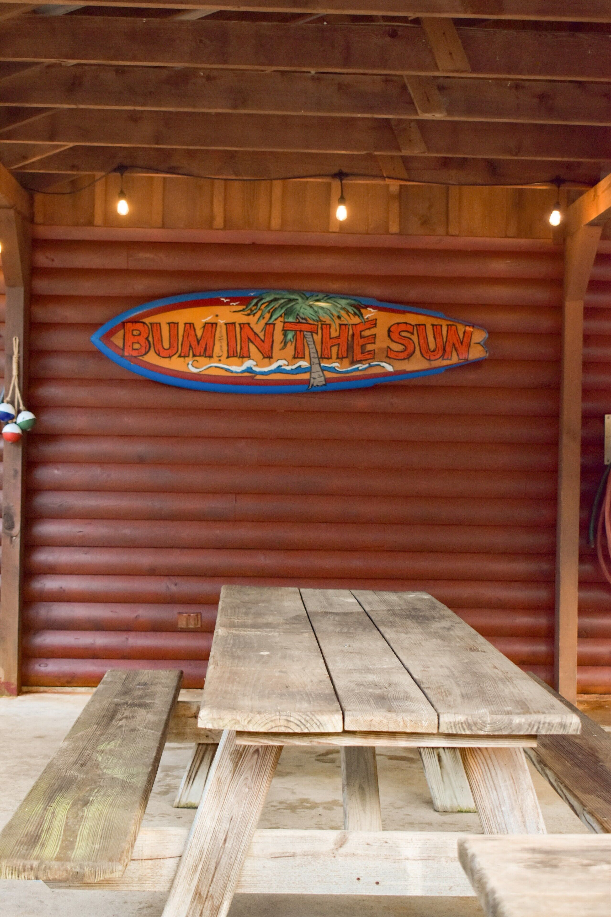 A wooden table and bench set against a red wooden wall with a sign that reads "BUMIN'THE SUN".