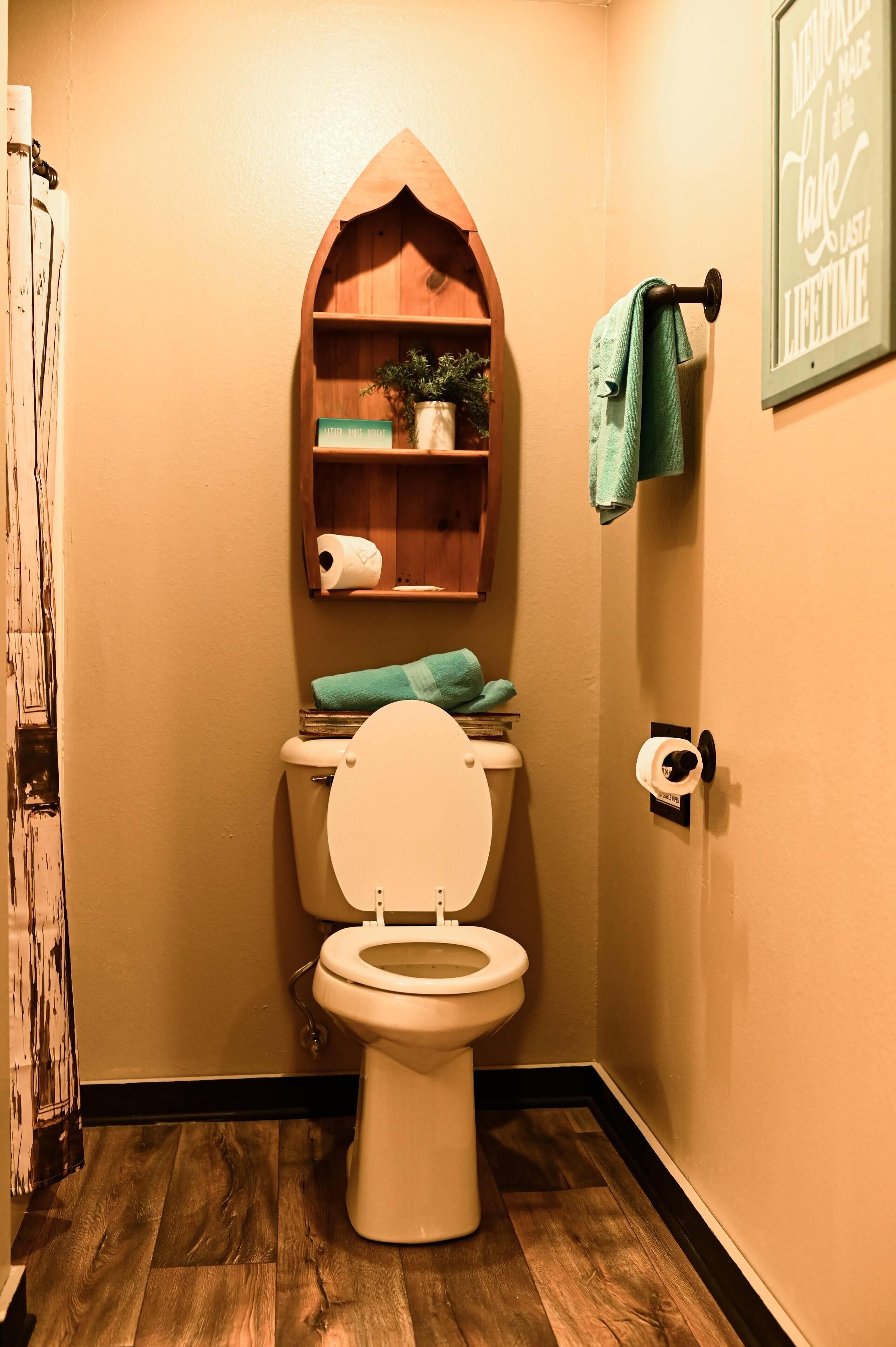 A bathroom with a wooden shelf above the toilet, a towel rack, and a framed sign on the wall.