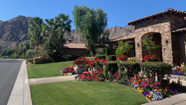A residential area with a stone house featuring an arched entrance, surrounded by well-maintained lawns and colorful flower beds. - Amazing 5 bedroom Indian Wells Country Club home with private backyard! (Indian Wells)
