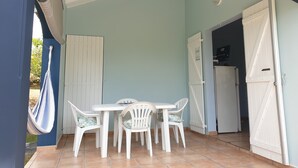 Une terrasse couverte avec une table et des chaises blanches, un hamac et une porte blanche donnant sur un espace cuisine.