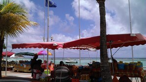 Un marché installé au bord de la plage, avec des étals sous des parasols rouges, un palmier et des bateaux en arrière-plan.
