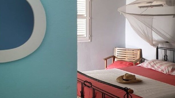 Une chambre à coucher avec un lit, une chaise en bois, un chapeau posé sur le lit et un ventilateur de plafond.