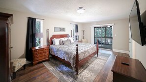 A bedroom with a large bed, a dresser, a nightstand, a TV, and a balcony with a view of trees.
