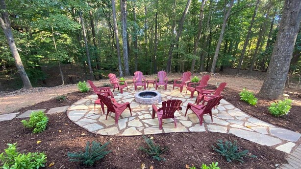 A circular outdoor seating area with Adirondack chairs and a fire pit, surrounded by a stone patio and small plants.