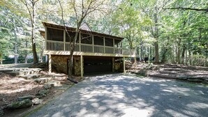 A rustic cabin with a wooden deck and a stone foundation, surrounded by a forested area.