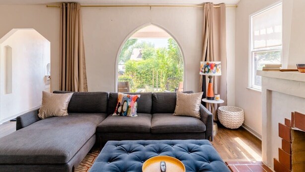 A modern living room with a sectional sofa, a tufted ottoman, a fireplace, and a view through a large arched window.