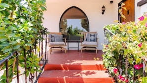 A covered outdoor seating area with two chairs and a table, surrounded by a wrought iron railing and climbing plants.