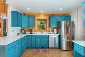 A kitchen with teal cabinets, a stainless steel refrigerator, and a window with a view of trees. - The Skybox - Voted #1 Cabin in Asheville (Mars Hill)