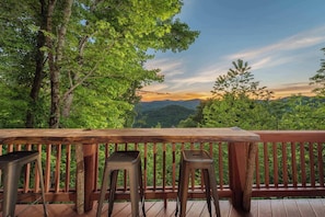 A wooden deck with a table and two stools, overlooking a forested landscape with distant mountains.