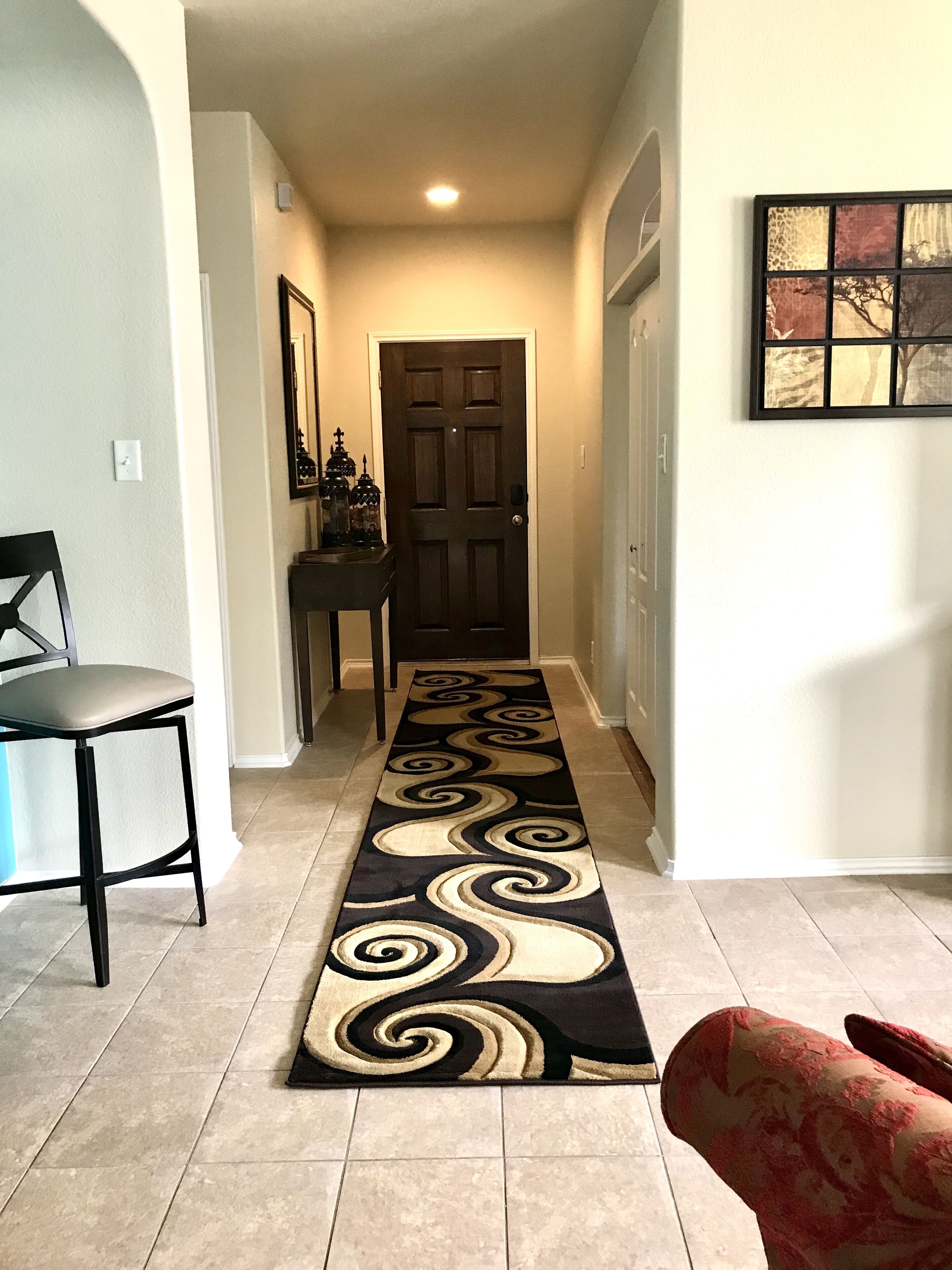 Un couloir avec un tapis à motifs, une porte, une chaise et une table ornée d’objets décoratifs.