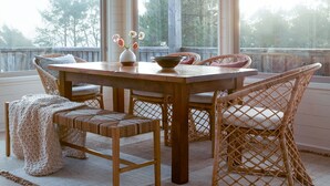 A dining area with a wooden table and wicker chairs, large windows, and a hanging light fixture. - Suil Na Mara - Modern Home with Mountain and Ocean Views (Tillamook County)