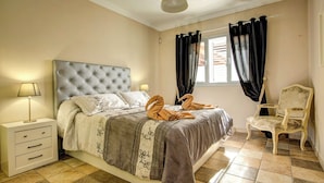 Une chambre à coucher avec une tête de lit capitonnée, un lit recouvert d’une couvre-lit à motifs gris et blancs, deux lampes de chevet, un fauteuil et une fenêtre avec des rideaux noirs.