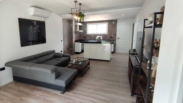 Uma sala de estar moderna com um sofá seccional cinza, uma mesa de centro de vidro e uma cozinha ao fundo.