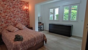 Um quarto com parede de tijolos aparentes, uma cama com roupa de cama estampada, uma mesinha de cabeceira, um penteado e uma janela com vista para o verde.