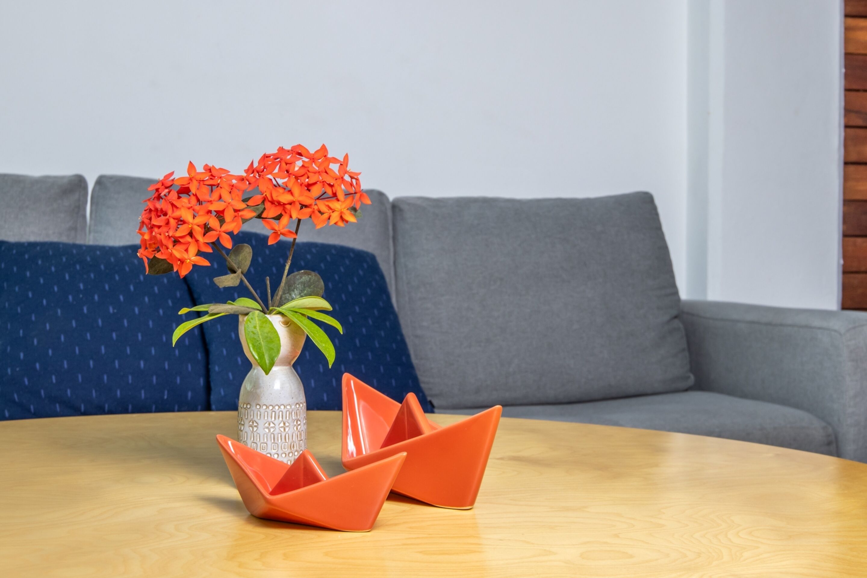 A round wooden table with a decorative vase containing orange flowers and green leaves, placed on a blue patterned sofa.