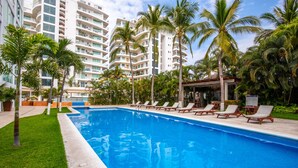 Una piscina con sillas de descanso, rodeada de palmeras y un edificio de varios pisos al fondo.