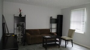 A living room with a brown sofa, a small table, a chair, a bookshelf, and a window with blinds.