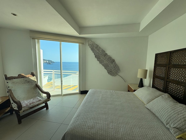 A hotel room with a bed, a chair, a large decorative leaf, a nightstand, and a view of the sea through a sliding door.