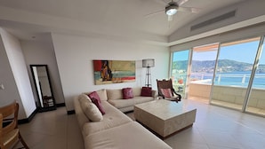A modern living room with a large sofa, a coffee table, and a chair. There is a painting on the wall and a large window with a view of the sea. - The Great Escape - Ixtapa - Penthouse on the Beach (Ixtapa Zihuatanejo)