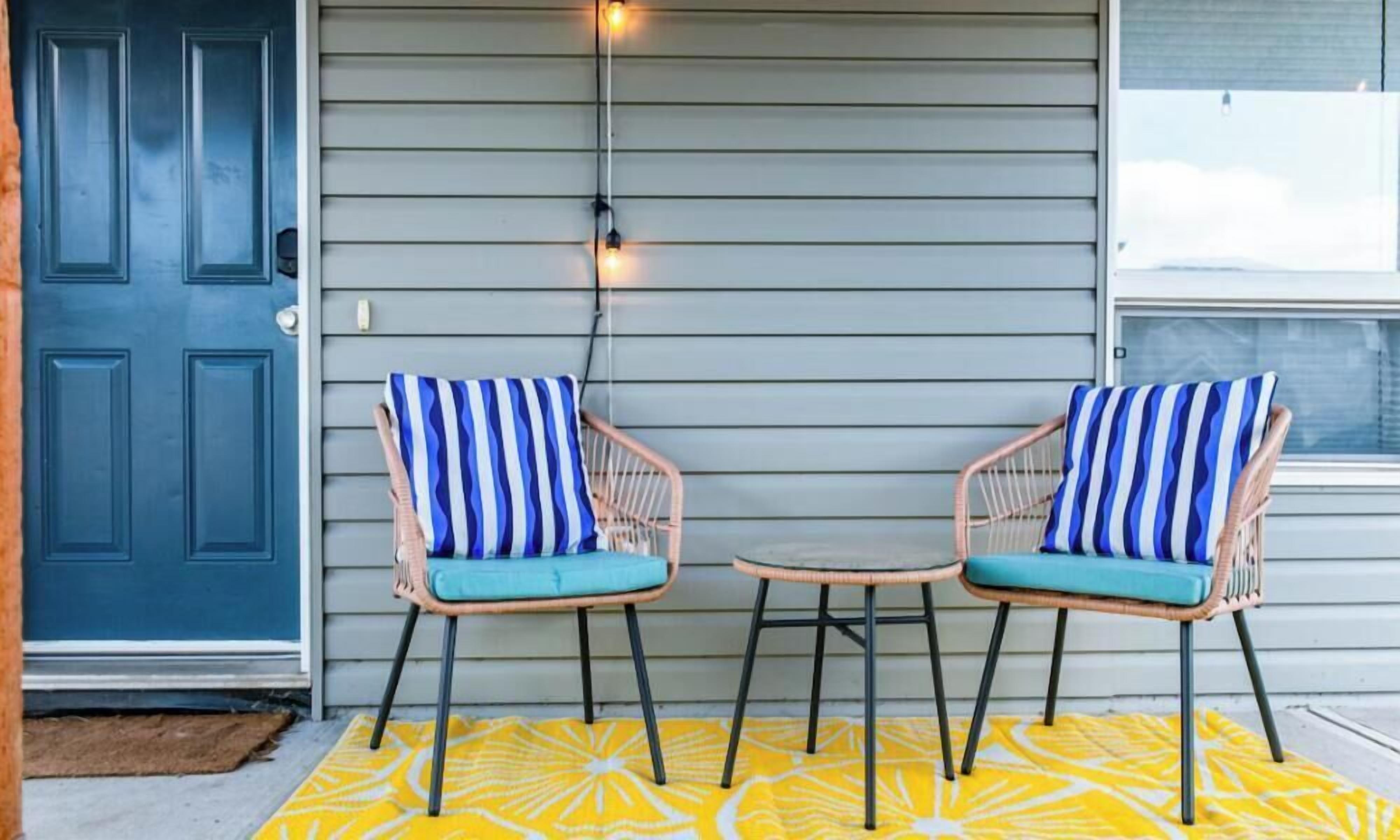 Two wicker chairs with striped cushions on a yellow patterned rug, a blue door, and a window with string lights.