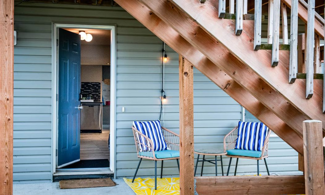 Een houten trap leidt naar een veranda met twee streepstoelen en een geel, patroonkleed.