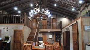 Un intérieur rustique avec un escalier en bois, un lustre orné de bois de cerf et un coin repas comprenant des chaises et une table en bois.