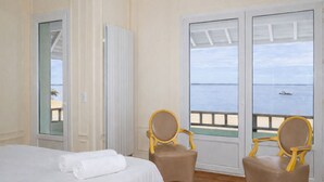 A hotel room with a bed, two chairs, a radiator, and a view of the sea.