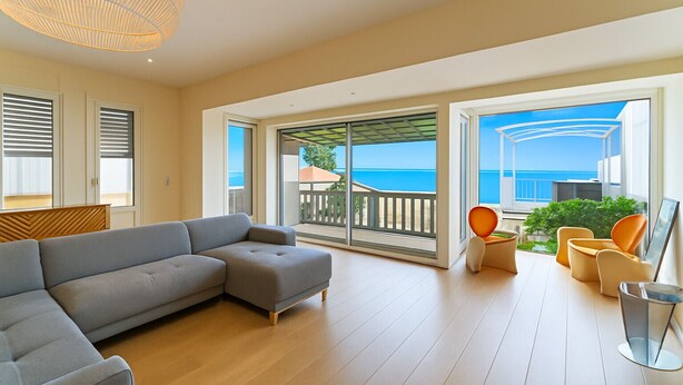 A spacious living room with a grey sectional sofa, a wooden floor, and a large sliding glass door leading to a balcony with a view of the sea.