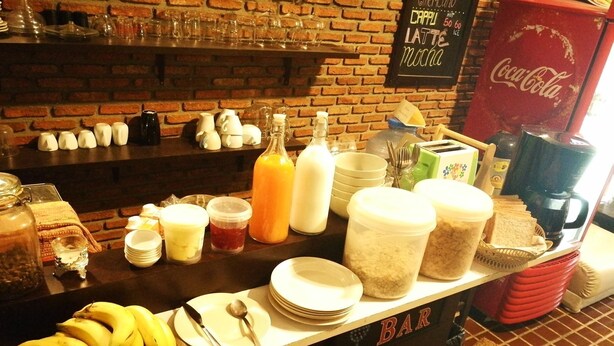A breakfast bar with various food items and beverages, including bananas, bread, and a Coca-Cola sign. - Hostel 16 (Bangkok)
