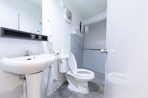 A bathroom with a white sink, toilet, and shower. There is a shelf above the sink and a window with blinds.