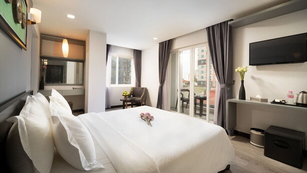 A hotel room with a large bed, a desk, a chair, a TV, and a view of the city through the window. - Le Cocon Boutique Hotel (Phnom Penh)