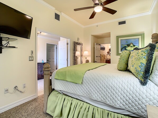 A bedroom with a large bed, a ceiling fan, a television mounted on the wall, and a door leading to another room.