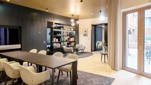 A modern living room with a dark wood dining table, beige chairs, a bookshelf, and a large flat-screen TV. - Chic Innere Stadt 1BR w/ Gym & Doorman, walk to Graben, by Blueground (Vienna)