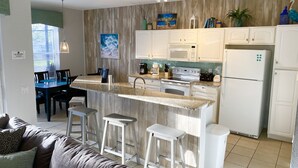 A modern kitchen with white cabinets, a granite countertop, and a built-in oven. There are bar stools, a dining table, and a couch in the adjacent living area. - Mickey’s Vacation Villa-Premium- Minutes from Disney World with private pool! (Kissimmee)