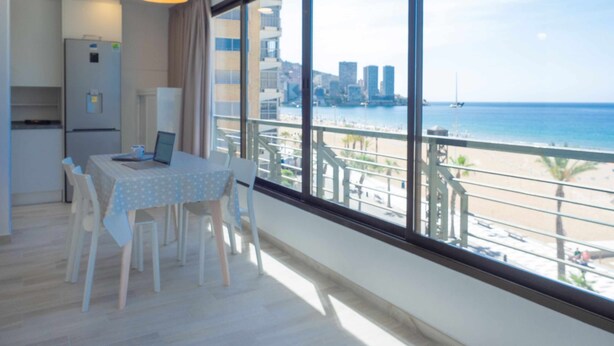 A modern apartment with a dining area, kitchen, and large windows overlooking a beach and cityscape. - Levante Beach Jazmines Ac (Benidorm)