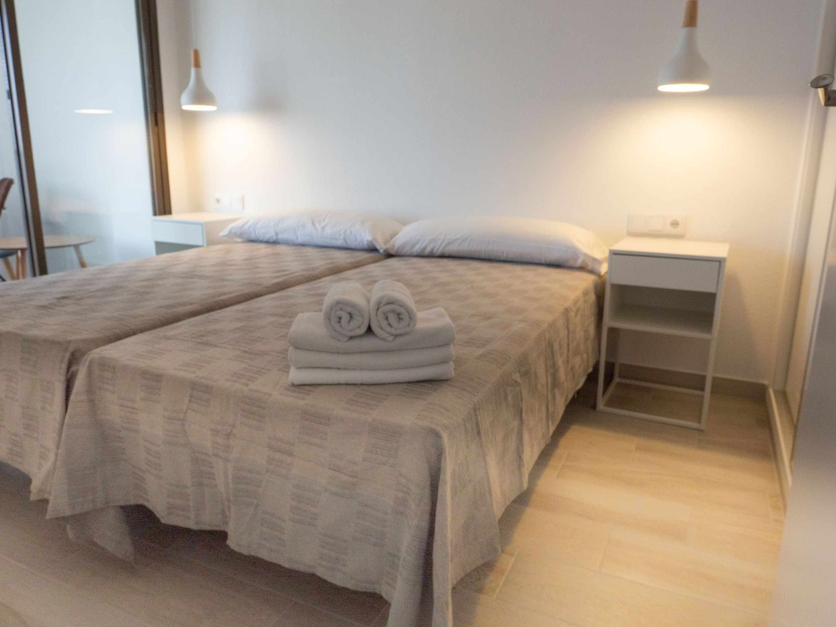 A double bed with a beige bedspread and two pillows, a small bedside table with a lamp, and a white nightstand with a shelf.