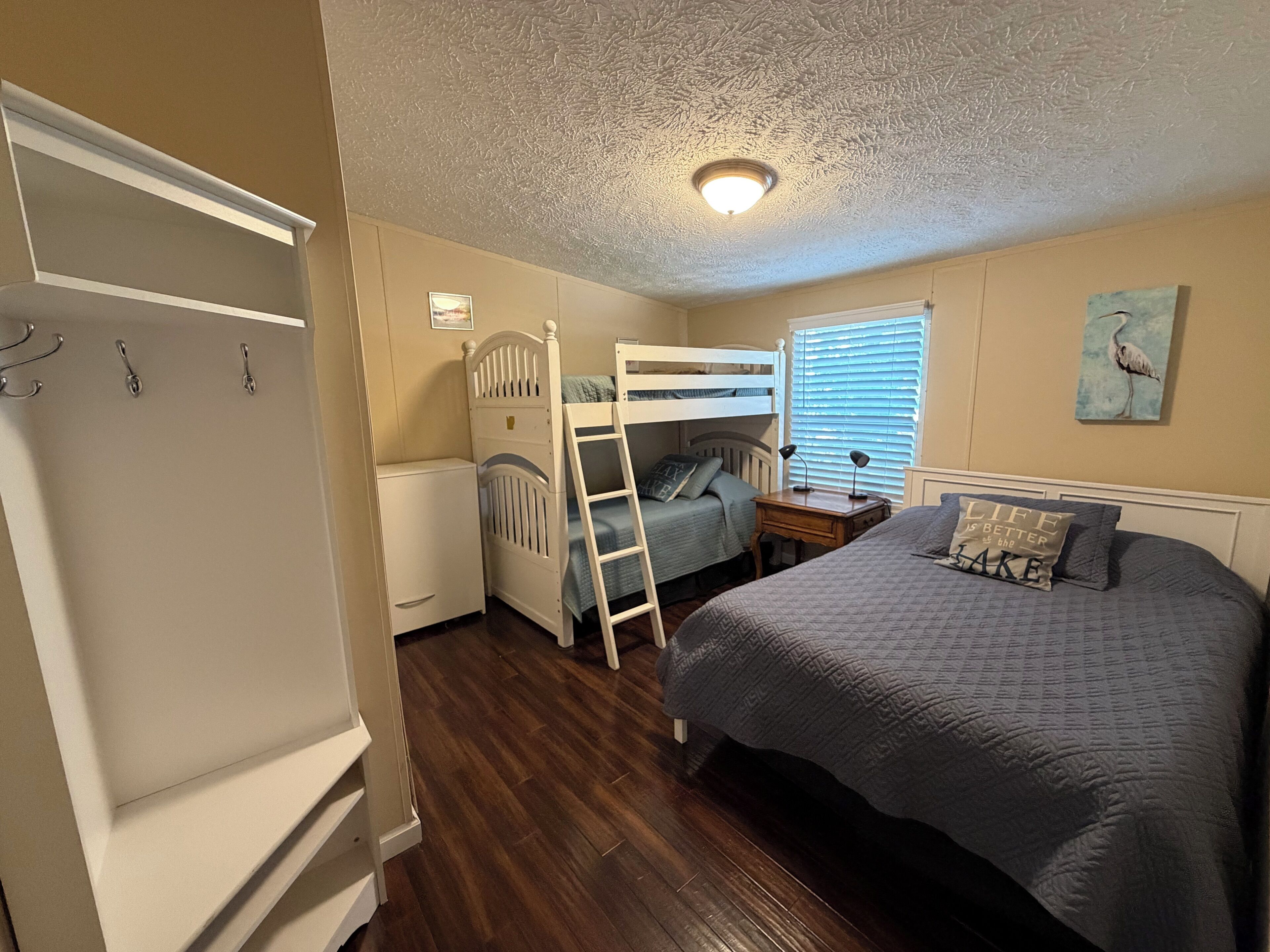 A bedroom with a bunk bed, a desk, a chair, a wardrobe, and a window with blinds.