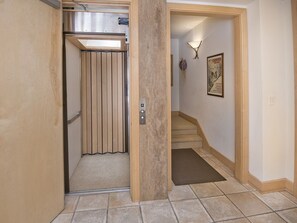 A hallway with a wooden door leading to an elevator, a staircase, and a wall-mounted light fixture.