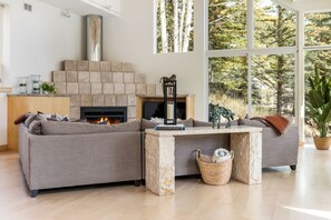 Una sala de estar moderna con chimenea de piedra, un sofá seccional, una mesa de centro y un ventanal con vista a árboles.