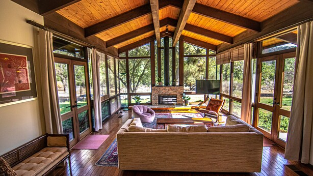 A spacious room with a wooden ceiling, large windows, a fireplace, and a sofa. - Kaweah River House ~ Private River Access (Three Rivers)
