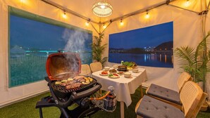 A tented outdoor dining area with a grill, table set with food, and views of a body of water. - Chuncheon El Dorado Kid's Pension (Chuncheon)