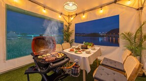 A tented outdoor dining area with a grill, table set with food, and views of a body of water. - Chuncheon El Dorado Kid's Pension (Chuncheon)