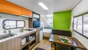 A compact kitchenette with a sink, a microwave, and a small refrigerator. There is a window with curtains, a television, and a small table with a magazine and a plant. - Gapeyong Unak Holiday Caravan (Gapyeong)