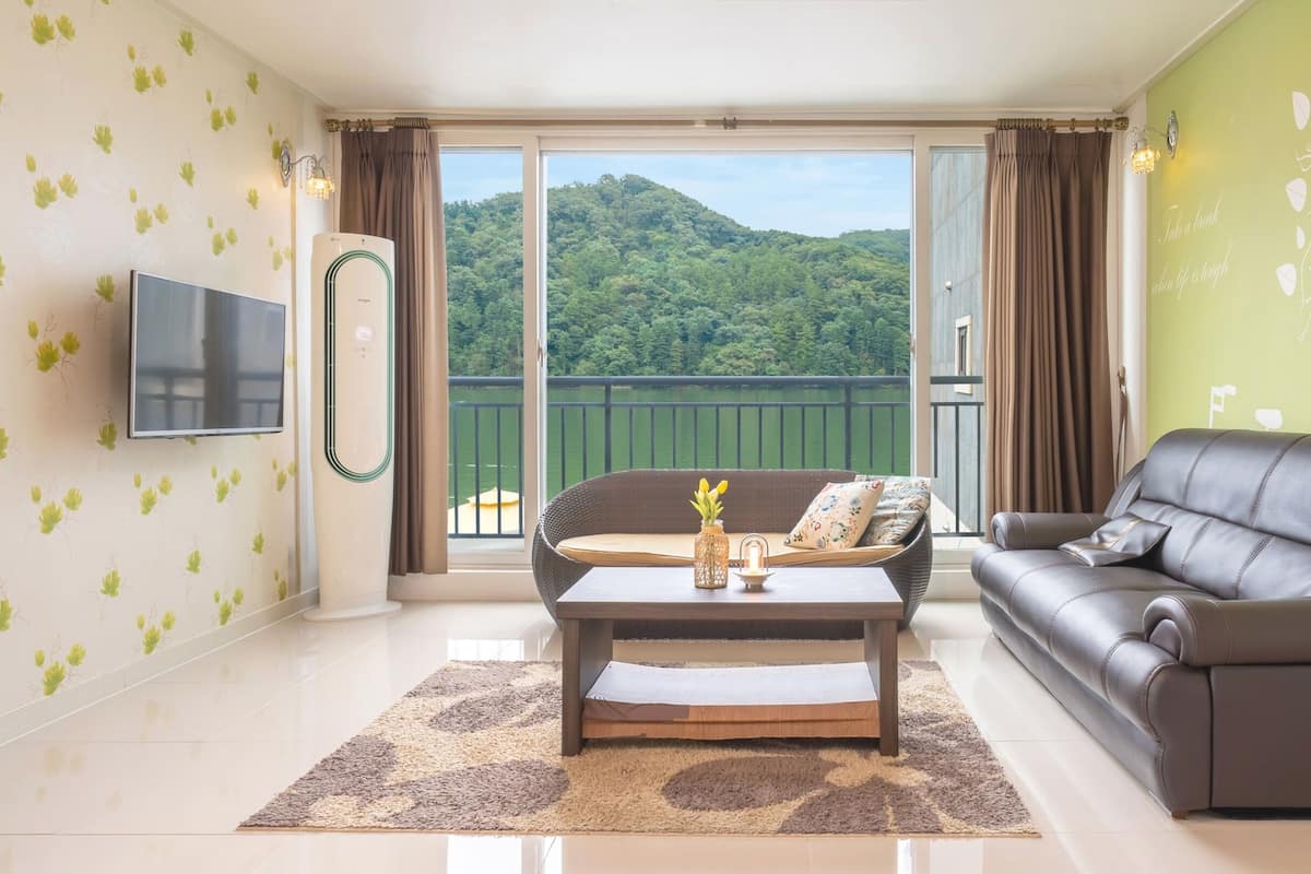A living room with a sofa, a coffee table, and a TV. There is a balcony with a view of a forest and a lake.