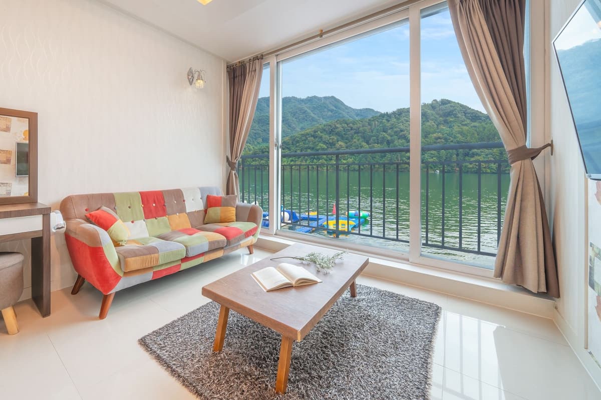 A modern living room with a large window, a colorful sofa, a small desk, and a view of a lake and mountains.